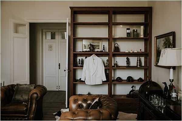 A groom's getting-ready detail shot taken indoors in what appears to be a well-furnished room within a historic property. A white dinner jacket or suit blazer hangs from a dark wood open bookshelf unit, which is decorated with an assortment of small figurines, decorative objects, and bottles on its shelves. In the foreground, a tufted brown leather Chesterfield armchair is partially visible, and a dark wood sideboard to the right holds several empty wine glasses and a table lamp with a white shade. The composition is a wide shot capturing the full room vignette, shot with a slightly moody, low-contrast edit.
