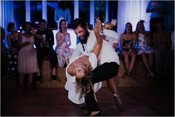 The couple is performing a dramatic dip move during their first dance at an indoor reception, with the groom holding the bride nearly upside down as she arches back with a wide smile. The groom is dressed in a white shirt and dark trousers, while the bride wears a white dress and has her hair loose. The reception space is lit with blue ambient uplighting, creating a moody atmosphere on the dance floor. Approximately 10-15 guests are visible in the background, seated and standing along the perimeter, reacting with visible excitement and applause. The shot is a wide portrait-style image capturing both the couple's movement and the crowd reaction.