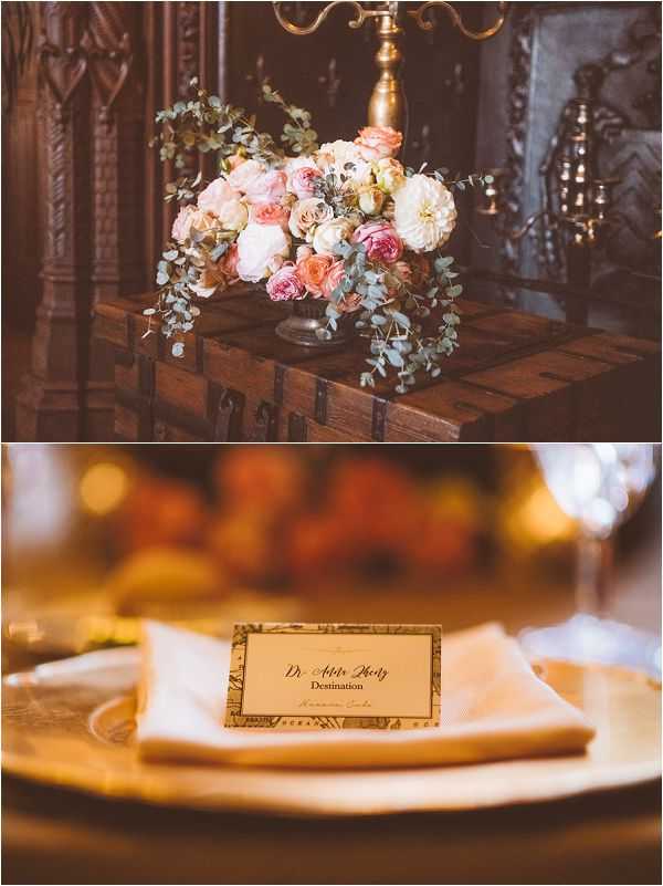A two-image detail collage from an indoor wedding reception with a classic, vintage-inspired aesthetic. The top image shows a low floral arrangement in a dark metal urn placed on an antique wooden trunk in front of an ornate carved fireplace; the arrangement features blush and coral garden roses, cream dahlias, and trailing eucalyptus. The bottom image is a close-up of a place setting featuring a gold charger plate, a folded ivory linen napkin, and a printed place card with a decorative map-style border reading 'Dr. Addie Khotz, Destination' — suggesting a travel-themed reception; a gold-tinted wine glass and warm ambient lighting are visible in the soft background.