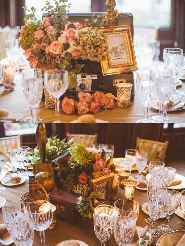 Close-up detail shots of indoor wedding reception table centerpieces styled with a vintage travel theme. The centerpieces feature open dark leather suitcases filled with coral and salmon roses, green hydrangeas, and eucalyptus foliage, accented with a small vintage film camera, a gold-framed table name card reading 'La Habana,' and gold mercury glass votive candle holders. Loose coral roses are scattered on the champagne-colored table linen around the suitcase base. The table settings include cut-crystal glassware in multiple sizes, white ceramic plates, and gold-toned bottle accents. The two-image composite shows the same tablescape from slightly different angles — one tighter close-up and one wider view showing the full table arrangement with guests' chairs visible in the background of an indoor reception space with warm candlelight.