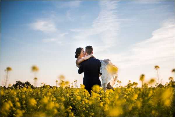A couple portrait shot outdoors in a field of bright yellow rapeseed flowers during golden hour. The groom, wearing a dark navy suit, is lifting the bride, who is dressed in a white gown, as they kiss. The composition is a wide shot with the couple silhouetted against a pale blue sky in the middle distance, framed by the vivid yellow blooms filling the foreground. The warm evening light creates a soft glow around the couple.