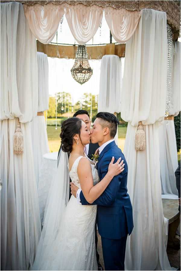 The bride and groom share their first kiss during an outdoor ceremony, framed by a draped altar structure adorned with cream and champagne-toned fabric panels tied back with tassel details and hanging crystal strands. A beaded chandelier hangs from the center of the canopy overhead, adding a classic decorative touch. The bride wears a fitted lace gown with a thin veil, while the groom wears a navy blue suit with a boutonniere; an officiant is partially visible standing behind them. The shot is a medium portrait taken straight-on, with a garden and warm late-afternoon light visible in the background through the draping.