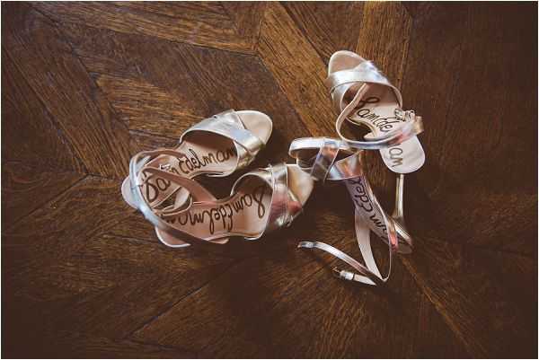 A close-up flat lay detail shot of a pair of silver strappy heeled sandals by Sam Edelman, placed on a dark herringbone parquet wood floor. The shoes are shown with ankle straps unwound, revealing the brand name written in cursive script on the tan insoles. The metallic silver straps catch the light against the warm tones of the wooden floor.