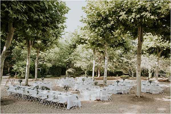 An outdoor wedding reception setup arranged beneath a canopy of tall trees on a gravel surface, with no guests present. Multiple long rectangular tables and a few round tables are dressed in white linens and lined with white folding chairs. Each table features low centerpieces of greenery and what appear to be small blooms in muted tones, placed at regular intervals along the table runners. The overall decor palette is white and green, with a clean, minimal aesthetic. Wide shot capturing the full layout of the reception space. Potential venue feature image.