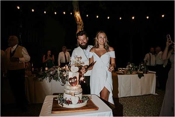 The bride and groom are cutting their wedding cake during an outdoor evening reception, surrounded by guests watching and photographing the moment. The bride wears an off-the-shoulder white wrap dress, and the groom is dressed in a white shirt. The naked-style tiered cake is decorated with greenery, dried florals, and sparklers, and topped with a personalized wooden laser-cut topper; it is displayed on a wooden board over a white linen-covered table. The venue is lit with warm string lights overhead, creating a dark, intimate atmosphere, and a white linen-covered head table is visible in the background with approximately a dozen guests gathered around. The shot is a medium portrait-style image taken at night with warm ambient lighting.
