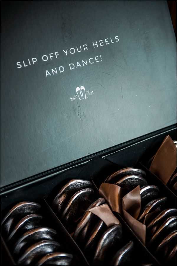 Close-up detail shot of a dark matte black gift box containing flat fold-up ballet slippers, a common wedding favor for guests to wear during the reception dancing. The inside lid of the box is printed in white text with the phrase 'SLIP OFF YOUR HEELS AND DANCE!' along with a small illustrated ballet slipper icon. The slippers inside appear to be dark bronze or black in color, individually packaged with brown tissue or satin wrapping dividers. The packaging design is modern and minimal, suggesting an intentional guest comfort favor detail.