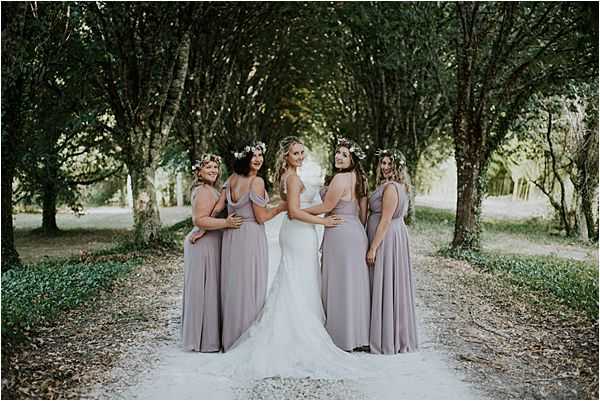 A bridal party portrait taken outdoors along a tree-lined pathway, with the bride surrounded by four bridesmaids. The bride wears a fitted white lace gown with a train, while all four bridesmaids wear floor-length dusty mauve/lilac chiffon dresses in varying neckline styles. Each bridesmaid wears a floral crown featuring small white flowers and greenery. The group is posed with their backs partially turned to the camera, with the bride and two bridesmaids looking back over their shoulders toward the lens. The styling has a relaxed boho aesthetic. Wide portrait shot with soft natural light filtering through the tree canopy overhead.