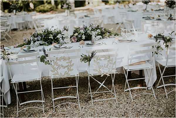 An outdoor wedding reception table setup photographed in a medium wide shot, showing a long rectangular dining table with a white linen tablecloth set on a gravel surface. Two white metal folding chairs in the foreground are decorated with laser-cut wooden wreath signs reading 'Groom' and 'Bride,' each adorned with small greenery sprigs. The table centerpiece features a lush garland of greenery with blush and mauve roses running along the center, accompanied by glassware and grey linen napkins at each place setting. Additional white folding chairs and guest tables are visible in the background, suggesting a garden or estate setting, with the overall decor palette being white, soft blush, and green in a classic French garden style.
