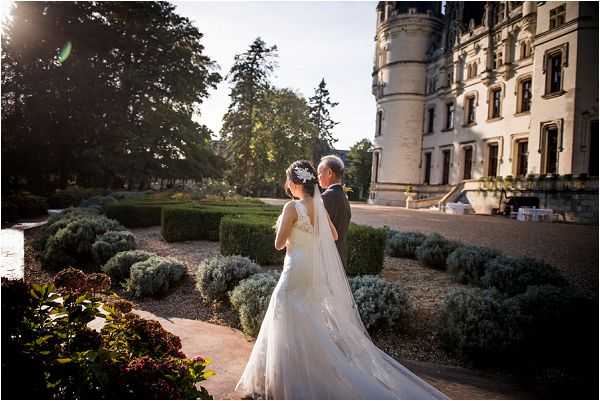 A bride and an older gentleman, likely her father, walk together through formal château gardens in a medium-wide shot taken from behind. The bride wears a white lace gown with a long cathedral-length veil and a floral hair accessory, while her companion wears a grey suit. The setting is the grounds of a large French château with ornate Renaissance-style stone architecture visible on the right side of the frame, and the formal garden features clipped hedgerows, lavender plantings, and gravel pathways. Warm late-afternoon golden light creates a lens flare effect and casts long shadows across the scene. Potential venue feature image.