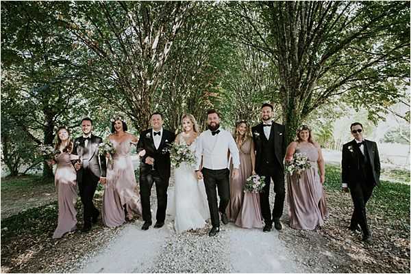 A bridal party of approximately ten people walks together along a gravel path lined with tall trees whose canopies form a natural arch overhead. The bride wears a white off-shoulder ballgown and carries a loose bouquet of white and greenery blooms, while the groom is dressed in a white dress shirt with black trousers and a black bow tie. The groomsmen wear classic black tuxedos with bow ties, and the bridesmaids are dressed in floor-length dusty mauve/blush satin gowns, some wearing floral crowns and carrying small bouquets. The group is mid-stride, walking toward the camera in a relaxed, candid style. The overall styling is classic with romantic boho touches, shot as a wide group portrait in an outdoor wooded setting.