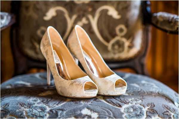 Close-up detail shot of a pair of bridal shoes displayed on an antique upholstered chair. The shoes are ivory peep-toe platform heels with a delicate lace or floral brocade fabric overlay and nude leather lining. They are placed on the blue-grey embroidered fabric seat of an ornate wooden chair with carved detailing visible in the background, suggesting a classic or vintage interior setting. The styling is classic and bridal, with the decorative chair serving as an intentional prop to complement the shoe design.