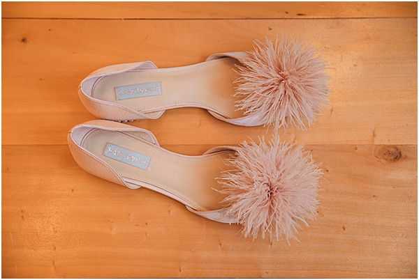 A flat lay detail shot of a pair of bridal flat shoes in blush pink, displayed on a light wood floor. The shoes feature a d'Orsay silhouette with large ostrich feather pom-pom embellishments in matching blush pink on the toe. A small blue brand label is visible inside each shoe, consistent with the 'something blue' bridal tradition. The composition is a top-down overhead shot showing both shoes side by side.