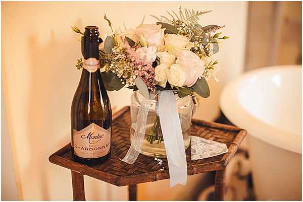 A detail close-up shot of a small wooden side table holding a bottle of Montuc Brut Chardonnay sparkling wine alongside a glass vase arranged with blush pink and ivory roses, eucalyptus, pink waxflower, and greenery, tied with a soft grey-blue ribbon. A piece of lace fabric is tucked beside the vase on the table surface. The warm indoor lighting and getting-ready setting suggest this is part of a bridal preparation scene. The overall styling palette is soft blush, ivory, and grey-blue with a romantic, classic feel.