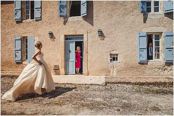 A bride stands outdoors in front of a traditional French stone building with powder blue shutters and a matching blue door, appearing to have a candid moment before the ceremony. She is wearing a full-skirted ivory ballgown with a structured bodice and has her hair styled in an updo. A woman in a fitted raspberry-red knee-length dress stands in the open doorway watching the bride, while a man in a suit is partially visible through one of the open windows to the right. The wide shot captures the rustic facade of what appears to be a French country property or small chateau, with the bride's voluminous dress train spread across the gravel courtyard. Potential venue feature image.