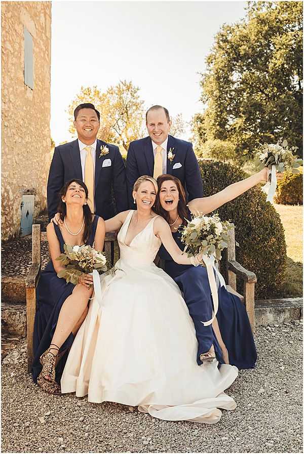 A bridal party portrait taken outdoors on a gravel courtyard in front of a stone building with pale blue-painted shutters, likely a French chateau or farmhouse. The bride sits center on a rustic wooden bench wearing an ivory ball gown with a V-neckline, flanked by two bridesmaids in navy blue floor-length dresses with pearl necklaces, all three laughing expressively; one bridesmaid raises her bouquet in the air. Two groomsmen stand behind in navy suits with yellow ties and boutonnières featuring small white flowers. The bouquets consist of ivory and cream blooms with eucalyptus and green foliage. The shot is a mid-range portrait with warm golden-hour lighting casting a warm tone across the group of five.