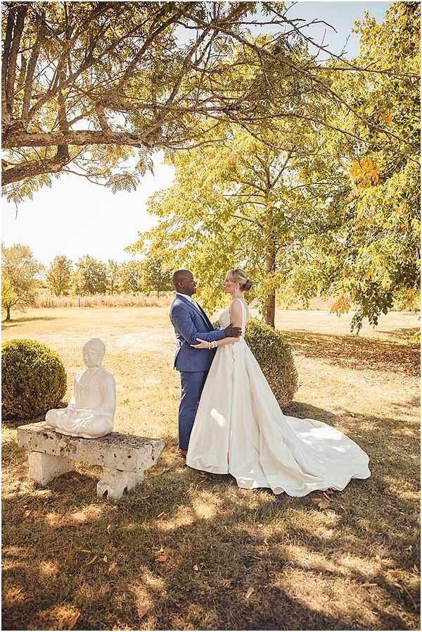 A couple portrait taken outdoors in the grounds of what appears to be a French country estate or chateau, with open fields visible in the background. The bride wears an ivory ball gown with a full, structured skirt and an extended train, with her hair styled in an updo, while the groom wears a fitted navy blue suit. The two face each other with arms around one another, standing beneath trees with warm golden-yellow autumn foliage. A white stone seated Buddha statue rests on a weathered stone bench to the left of the couple, flanked by trimmed boxwood spheres, suggesting a classic French garden setting. The shot is a full-length portrait taken in warm autumn afternoon light.