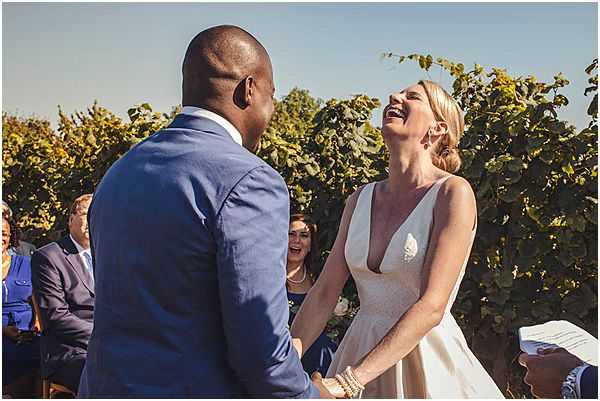 An outdoor wedding ceremony taking place in front of a dense green vineyard backdrop, captured during what appears to be a bright, sunny day. The bride, wearing a white deep V-neck dress with a leaf or feather detail at the bodice, is laughing openly while holding hands with the groom, who is dressed in a blue suit and faces away from the camera. A small group of seated guests is visible in the background, including a woman in a navy dress who is also smiling and reacting to the moment. The composition is a candid mid-shot focusing on the couple's interaction during the ceremony exchange.