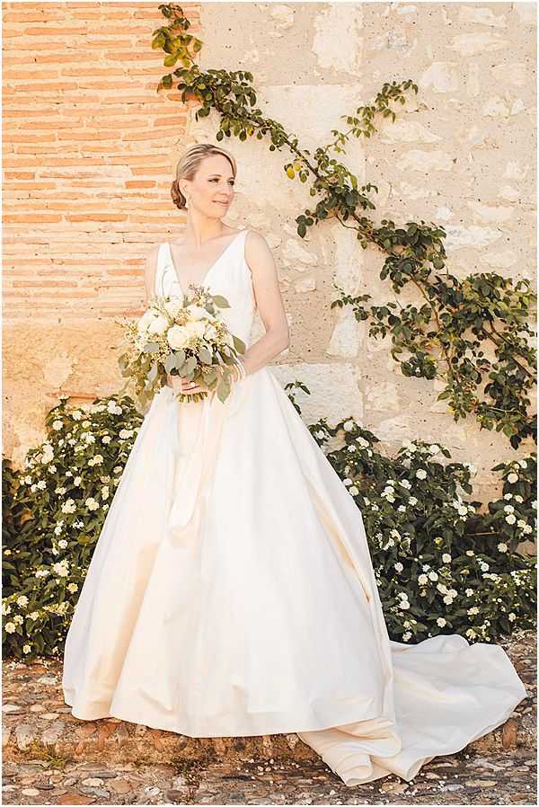 A bridal portrait of a single bride standing outdoors against an aged stone and brick wall. She wears an ivory satin ball gown with a V-neckline, a full skirt, and a trailing train, and holds a bouquet of cream roses, white blooms, and trailing eucalyptus and greenery. Her hair is styled in a low updo. The composition is a full-length portrait shot with warm natural light, giving the image a golden tone.