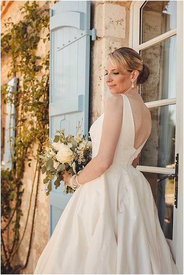 A bridal portrait taken outdoors against the facade of a French stone building featuring pale blue wooden shutters and a white-framed window. The bride stands in three-quarter profile, glancing back over her shoulder toward the camera, wearing an ivory ball gown with a low open back and full skirt. Her hair is styled in a low chignon, and she wears drop earrings and pearl bracelets. She holds a bouquet of cream garden roses, eucalyptus, and small white filler flowers with trailing greenery. The warm golden light suggests late afternoon or early evening. The composition is a medium portrait shot with a shallow depth of field, keeping the bride sharp against the softly blurred architectural background.