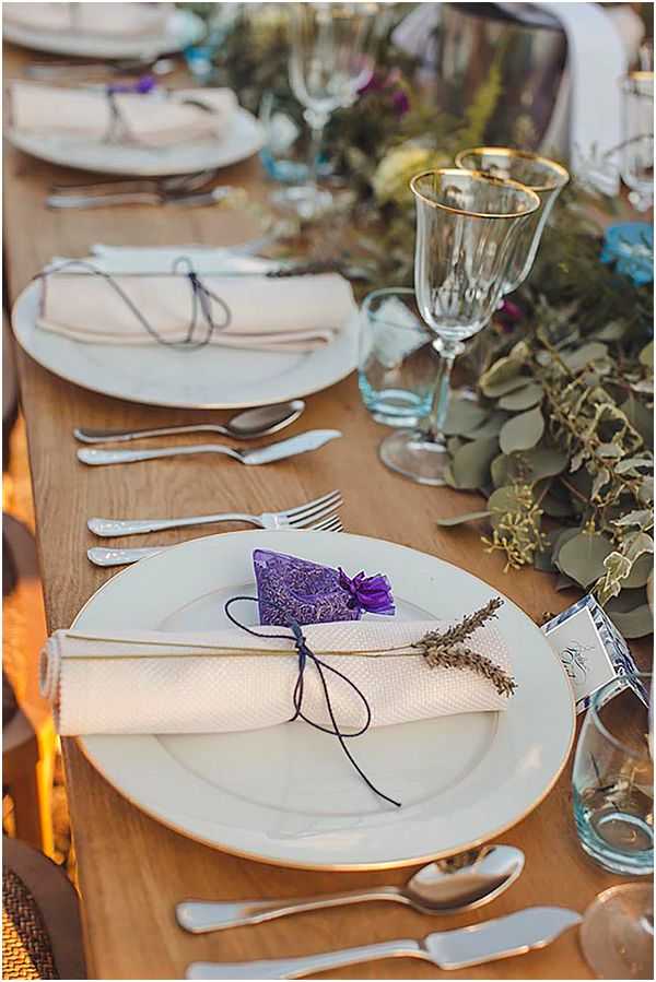 Close-up detail shot of a wedding reception table setting on a bare wooden farm table. The foreground place setting features a white plate with a cream linen napkin rolled and tied with dark twine, topped with a small purple lavender sachet and a sprig of dried lavender as a guest favor. Silver cutlery is arranged on either side of the plate, and glassware includes a cut-crystal goblet with a gold rim and a blue-tinted water glass. A lush garland runner of eucalyptus, greenery, and small purple and yellow blooms runs the length of the table. The styling is rustic-Provençal with a lavender and eucalyptus color palette.