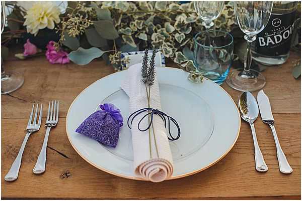 Close-up detail shot of a single place setting on a rustic wooden table. A white plate with a gold rim holds a rolled linen napkin tied with dark navy cord and two dried lavender stems, accompanied by a small purple organza sachet filled with lavender as a guest favor. Silver cutlery — two forks on the left and a knife and spoon on the right — flank the plate. In the background, a floral centerpiece features eucalyptus greenery, ivory dahlias, and purple and pink blooms, alongside a Badoit water bottle and clear glassware including a wine glass and champagne flute. The overall decor palette combines navy, purple, and natural tones with a Provençal-inspired styling theme.