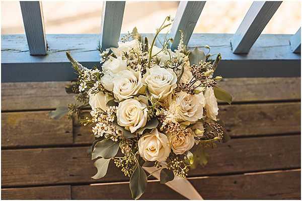 Close-up detail shot of a bridal bouquet resting on a weathered wooden surface in front of painted blue-grey lattice panels. The bouquet is composed of cream and blush roses in various stages of bloom, white spray flowers, dusty sage eucalyptus leaves, and small dried berry accents in muted brown tones. The overall palette is soft ivory, champagne, and sage green with a rustic, organic styling. Pale blush ribbon trails from the bouquet stems onto the wooden planks.