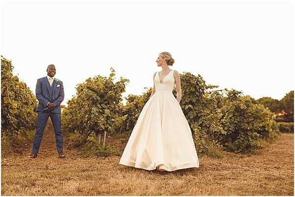 A couple portrait taken outdoors in a vineyard during golden hour light. The bride wears a full-skirted ivory satin ball gown with a deep V-neckline and has her hair styled in an updo; she stands in the foreground with her hands in the skirt pockets, turning slightly toward the groom. The groom stands several feet behind her wearing a navy three-piece suit with a white pocket square and a boutonniere. The composition is a wide shot with rows of lush green grapevines stretching across the background, and the warm late-day light gives the scene a golden tone. The overall styling is classic and clean.