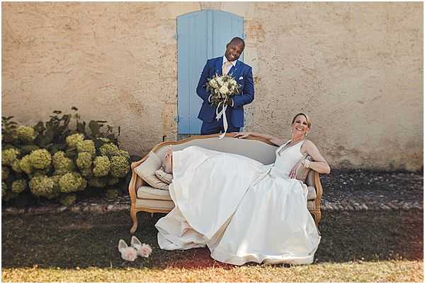 A couple poses outdoors against a weathered stone wall featuring a pale blue wooden door, with the bride seated on a gold-framed antique loveseat and the groom standing on it behind her. The bride wears a full-skirted white ballgown with a V-neckline, her heels discarded on the grass nearby, and she laughs toward the camera in a relaxed pose. The groom wears a cobalt blue suit with a white dress shirt and holds the bridal bouquet, which features cream and white blooms with trailing ribbons. The styling has a relaxed, classic French countryside feel, and the shot is a medium-wide portrait capturing both figures and the decorative seating prop.