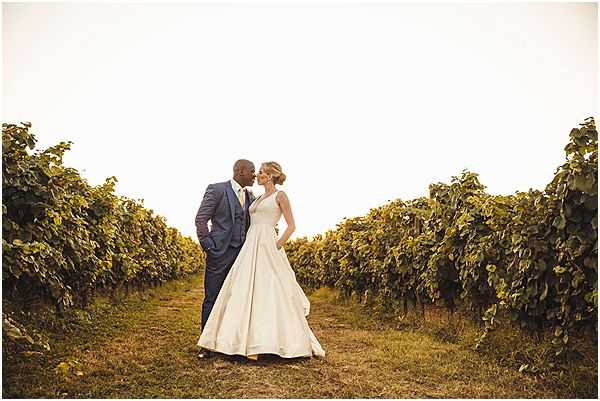 A couple portrait taken outdoors in a vineyard, with rows of grapevines extending symmetrically on either side of the couple. The groom wears a fitted navy blue three-piece suit, and the bride wears a full-skirted ivory satin ball gown with a structured bodice and an updo hairstyle. The two face each other closely in an intimate moment. The image is shot in warm golden-hour light, giving the scene an amber tone. The composition is a wide shot with the couple centered between the vine rows, creating strong leading lines.
