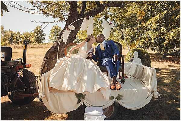 The bride and groom share a kiss while seated outdoors beneath a large tree in a styled vignette setting. The bride wears a voluminous ivory/cream ball gown with a full skirt, while the groom is dressed in a cobalt blue suit with brown leather oxford shoes and a light blue pocket square. They are seated in vintage-style chairs draped with white fabric, accompanied by a white classical bust sculpture, creating an eclectic, artistic aesthetic. A hoop arch decorated with white fabric streamers and small floral accents frames them from above, and a vintage tractor is partially visible to the left; the overall styling blends boho and vintage-romantic elements. Wide portrait shot taken in bright midday sunlight in an open rural landscape.