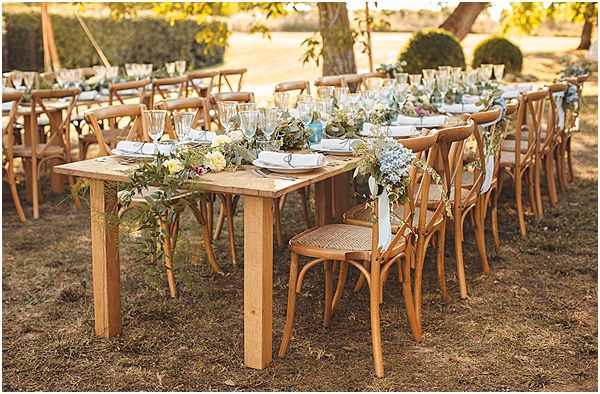 An outdoor wedding reception table setup photographed in a wide shot within a garden or parkland setting. The long wooden farm-style tables are surrounded by natural wood cross-back chairs, set with white linen napkins, glassware including wine and water glasses, and what appear to be charger plates. A lush floral garland runner runs along the center of the table, featuring soft blue hydrangeas, cream and pale yellow roses, and trailing eucalyptus and greenery, with small blue glass bud vases interspersed along the runner. The overall decor palette is natural and rustic, combining warm honey-toned wood furniture with a muted blue, cream, and green floral scheme. The styling is relaxed rustic with a garden-party feel, and the warm golden light suggests late afternoon sun.