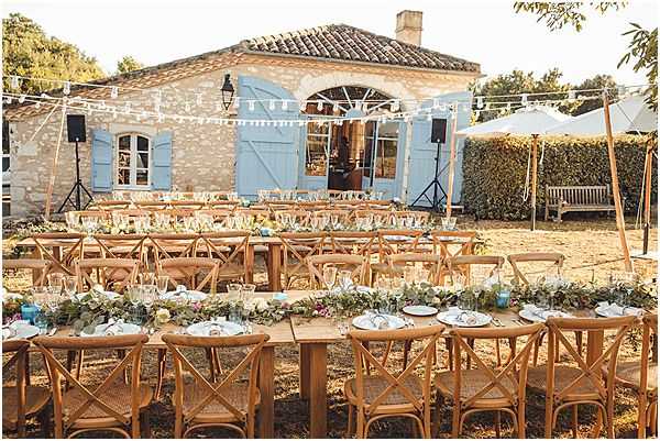 An outdoor wedding reception setup in the courtyard of a stone Provençal mas or farmhouse with pale blue shutters and terracotta roof tiles. Multiple long wooden farm tables are arranged in rows, lined with natural cross-back wooden chairs and dressed with lush greenery garland runners featuring white and soft-toned blooms, glassware, and white plate settings. String fairy lights and white triangle bunting are strung between posts above the dining area, and large white market umbrellas are visible to the right. The overall styling is relaxed rustic-Mediterranean with warm natural wood tones, greenery-forward florals, and a casual open-air feel. Wide establishing shot capturing the full reception layout. Potential venue feature image.