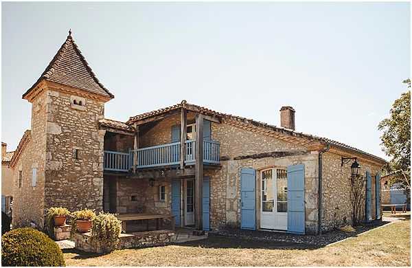 Wide exterior shot of a French countryside stone building featuring a distinctive pointed tower with a tiled conical roof, pale blue shutters, blue-painted doors and balcony railings, and a covered upper terrace. The rustic stone architecture is characteristic of the Southwest French region, with terracotta roof tiles and arched doorways. No people are visible in the frame. Potential venue feature image.