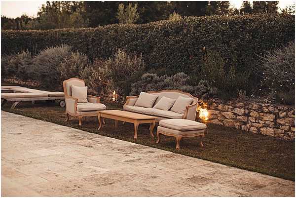 An outdoor cocktail hour or reception lounge area photographed at dusk, with no people visible. The seating arrangement consists of a French Louis-style sofa, a matching armchair, an ottoman, and a low wooden coffee table, all upholstered in neutral linen or beige fabric with carved wooden frames in a natural finish. Small candle lanterns placed on the ground provide warm ambient lighting along a low stone retaining wall. The setting is on a manicured lawn adjacent to a stone-paved terrace, with a dense hedge backdrop and lavender plantings contributing to a Provençal garden atmosphere. The overall decor style is classic French country. Wide establishing shot. Potential venue feature image.