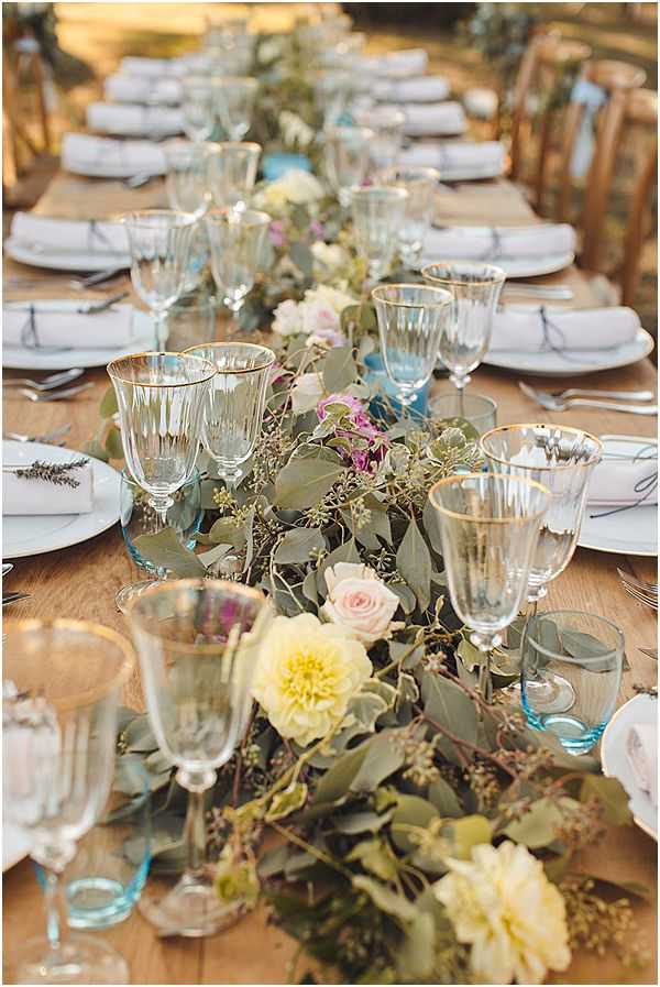 Close-up detail shot of a long outdoor reception dining table set for a wedding. The table is a raw wood farm-style surface dressed with a lush greenery runner composed of eucalyptus and trailing foliage, interspersed with yellow dahlias, blush pink roses, and small purple wildflowers. Place settings include white plates with white linen napkins, and each setting features a mix of mismatched vintage-style glassware including clear crystal wine glasses with gold rims and soft blue-tinted glass goblets, giving a collected, boho aesthetic. Cutlery is laid on either side of the plates and small sprigs of lavender are visible as accents. The overall decor palette is soft and muted — ivory, blush, pale yellow, sage green, and powder blue — consistent with a relaxed, garden-style boho wedding.