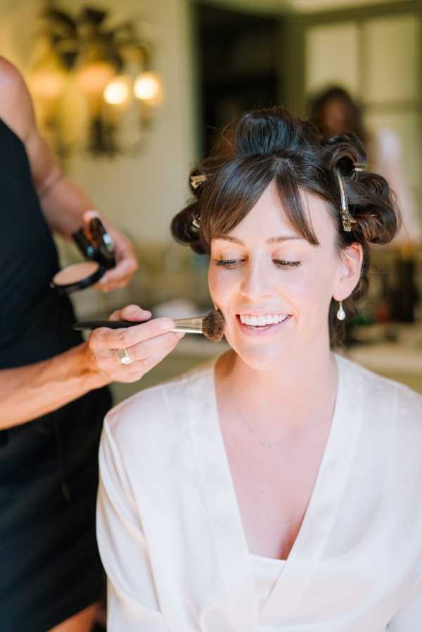 A getting-ready scene showing a bride seated while a makeup artist applies powder to her cheek with a brush. The bride has dark brown hair set in large velcro rollers and clip sections, and is wearing a white satin robe. She is smiling with eyes slightly closed and wearing delicate pearl drop earrings and a fine necklace. The setting appears to be an indoor dressing room or bathroom, with warm wall sconce lighting visible in the background. The shot is a close-up portrait taken at medium distance, with the makeup artist's hands and a compact powder case visible in the foreground.