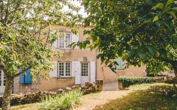 Wide exterior shot of a traditional French country house with warm sandy-beige rendered walls, white-framed windows, and pale blue shutters. The two-storey property features classic provincial architecture with a ground-floor glass-paned door and a small balcony railing visible on the upper level. The image is framed through mature trees in the foreground, with a gravel pathway and low stone borders visible on the grounds. No people are present in the shot. Potential venue feature image.