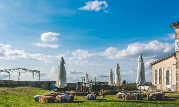 A wide shot of an outdoor wedding cocktail hour or reception setup on the grounds of what appears to be a French stone chateau or domaine. The seating area features hay bales topped with fabric cushions or blankets in muted tones arranged across a lawn, with several closed white market umbrellas providing shade. A bar or DJ setup is visible in the background with a small group of people gathered around it, and a greenery-covered pergola or arbor structure stands to the left. The styling is rustic and relaxed, consistent with a countryside or vineyard wedding aesthetic. The venue building, constructed of pale stone with tall windows, is partially visible on the right edge of the frame. Potential venue feature image.