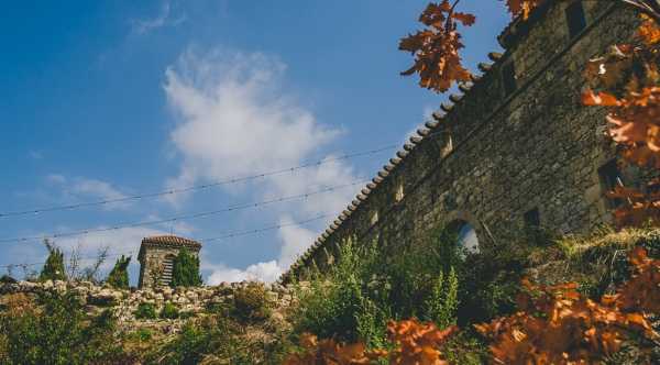 A wide-angle architectural shot looking upward at the exterior stone walls and tower of what appears to be a historic French chateau or fortified property, taken in autumn with orange-brown foliage visible in the foreground. No people are present in the image. String lights are strung between the structures, suggesting event use. The composition emphasizes the aged stonework, arched window openings, and a small stone turret in the background. Potential venue feature image.