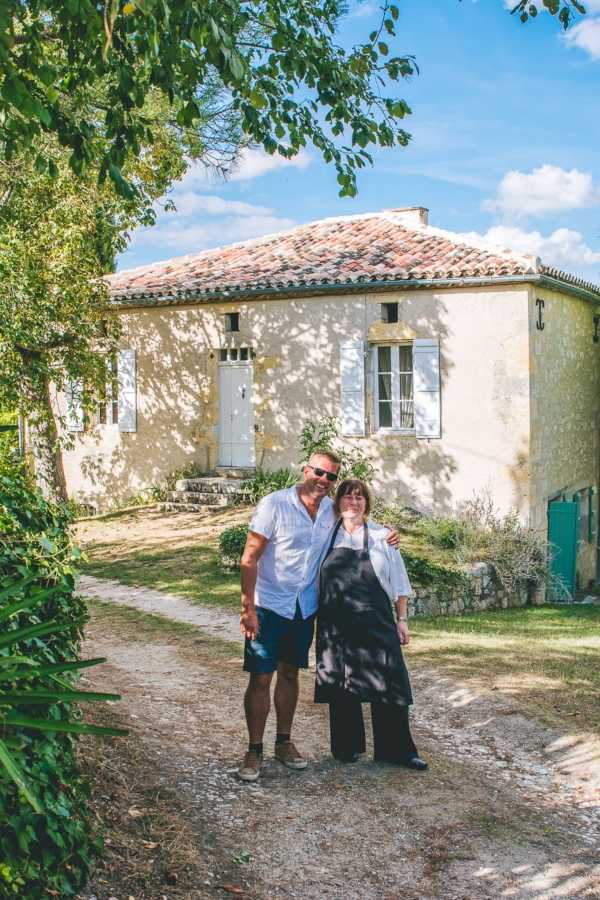 A couple poses together on a gravel path in front of a traditional French stone building with terracotta roof tiles, white shutters, and a white door. The man wears a white linen shirt, navy shorts, and tan shoes with sunglasses, while the woman wears a black long-sleeve top and wide-leg black trousers. They stand close together with his arm around her shoulders, smiling at the camera. The shot is a medium portrait taken outdoors in bright daytime light, with dappled tree shadows falling across the building facade and a teal-painted garage door visible to the right.