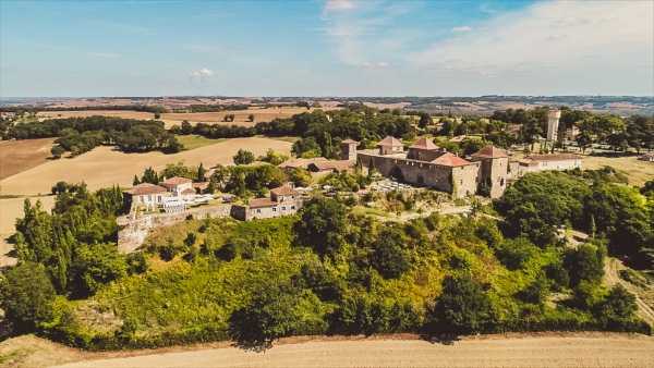 Aerial wide shot of a French château complex situated on a hilltop, surrounded by mature trees and rolling countryside. The property features multiple stone buildings with terracotta-tiled roofs, a tower, and what appears to be an outdoor terrace or event space visible on the right side of the complex. The surrounding landscape shows open agricultural fields extending to the horizon. Potential venue feature image.