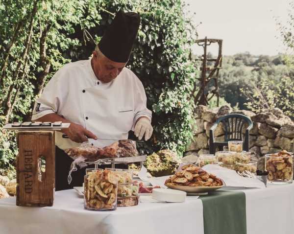 A chef in a white double-breasted jacket and black toque is carving a large roast at an outdoor cocktail hour or buffet station. The table is covered with a white linen and styled with a sage green table runner, and is laid out with multiple food items including glass jars filled with dried fruit or chips, a wooden crate branded with 'Hédard,' platters of toasted bread slices, and various other appetizers. The setting is an outdoor terrace or garden area with a rustic stone wall and dense green foliage in the background. The shot is a medium wide composition capturing the full spread of the food station and the chef at work.