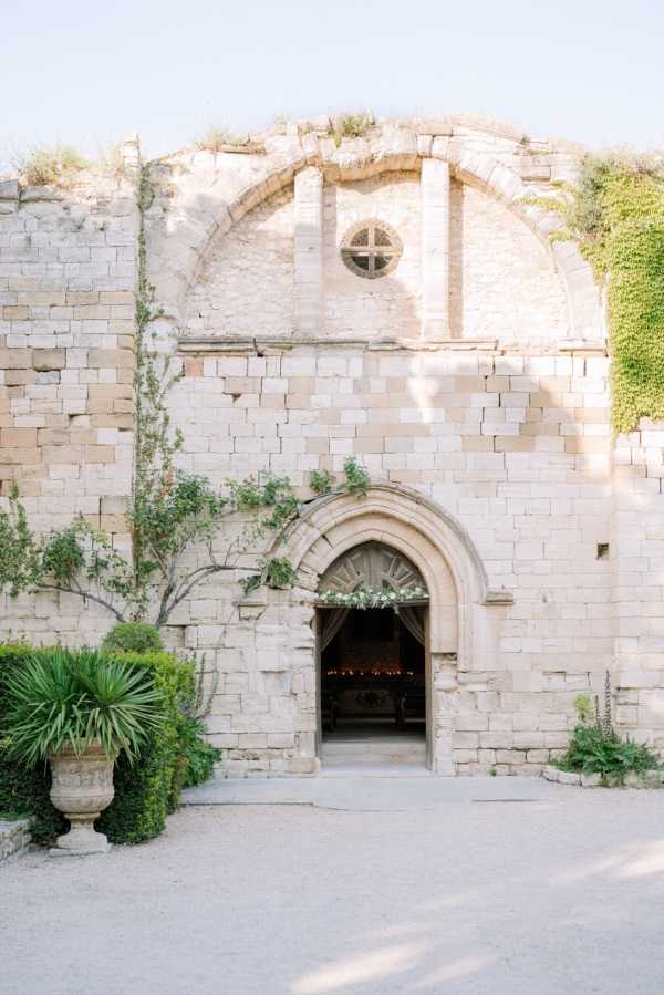 A wide shot of the exterior facade of an ancient stone chapel or abbey with a rounded Gothic arch doorway decorated with a greenery and white floral installation across the top. The interior of the chapel is visible through the open doorway, with warm candlelight or string lights glowing inside. The facade features a circular rose window, climbing vines, and partially ruined upper walls. Two large stone urns with trimmed boxwood and palm-like plants flank the entrance on a gravel courtyard. No people are visible in the frame. The overall decor style is classic and organic, with the greenery arch complementing the historic architecture. Potential venue feature image.