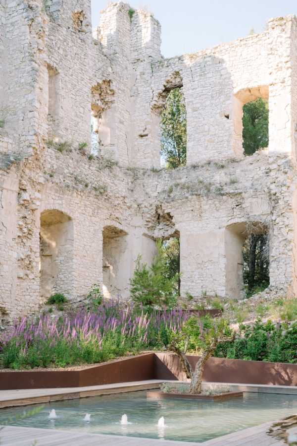 A venue detail shot showing the interior courtyard of a ruined stone castle or château, with open-air roofless walls featuring large arched openings across multiple levels. In the foreground, a rectangular reflecting pool with small fountain jets is framed by Corten steel raised planters filled with purple and pink flowering plants, likely salvia and foxglove, along with green shrubs and a small tree. No people are visible in the image. The overall styling aesthetic blends historic ruins with modern landscaping design. Potential venue feature image.