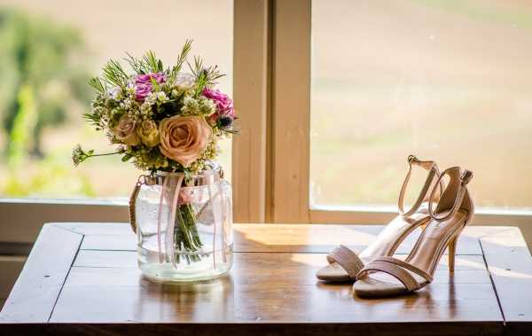 A bridal detail flat lay showing a bridal bouquet in a glass jar vase and a pair of nude strappy stiletto heels placed on a rustic wooden table in front of a window. The bouquet features peach and blush roses, pink wildflowers, thistle, baby's breath, and pine foliage, tied with a pink ribbon. The heels are a champagne-nude tone with ankle straps and gold hardware. Natural light comes through the window, with a soft green outdoor background visible behind the glass.