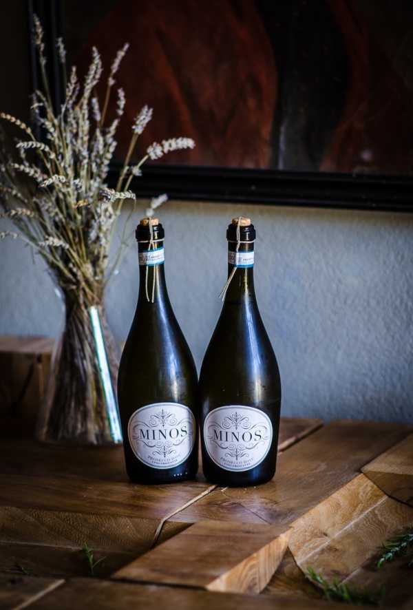 Close-up detail shot of two bottles of Minos Prosecco displayed on a rustic wooden surface, likely a welcome table or bar setup at a wedding reception. The bottles feature cream and black oval labels with decorative typography and small gold tags tied around their necks. In the background, a glass vase holds dried lavender and wheat-toned grasses, alongside a dark-framed artwork, contributing to a rustic, warm-toned decor scheme. Small sprigs of fresh rosemary are scattered on the wooden board in the foreground.