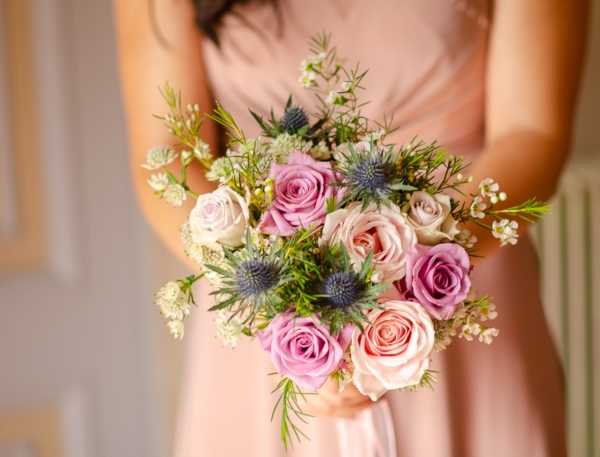 A close-up detail shot of a bridesmaid holding a hand-tied bouquet composed of blush and mauve roses, blue-grey eryngium (sea holly) thistles, white waxflower, Queen Anne's lace, and fine green foliage. The bridesmaid is wearing a blush pink dress, visible from the torso down in soft focus behind the bouquet. The setting appears to be indoors, likely a getting-ready or portrait moment, with a neutral-toned interior wall in the background. The overall styling palette is romantic and garden-inspired, mixing soft pinks with contrasting textural blue-grey thistle accents.