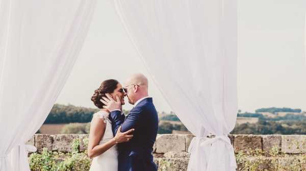 A bride and groom share a kiss or near-kiss during what appears to be an outdoor ceremony or post-ceremony portrait on a stone terrace or balustrade, with a rolling countryside landscape visible in the background. The couple is framed by a white fabric draping arch or canopy structure tied at the sides, creating a soft, airy frame around them. The bride wears a white lace cap-sleeve gown with her dark hair in an updo, while the groom is dressed in a navy blue suit; he is cupping her face gently with one hand. The shot is a medium portrait composition taken from a slight distance, with the white drapes and open sky giving the image a light, airy feel.