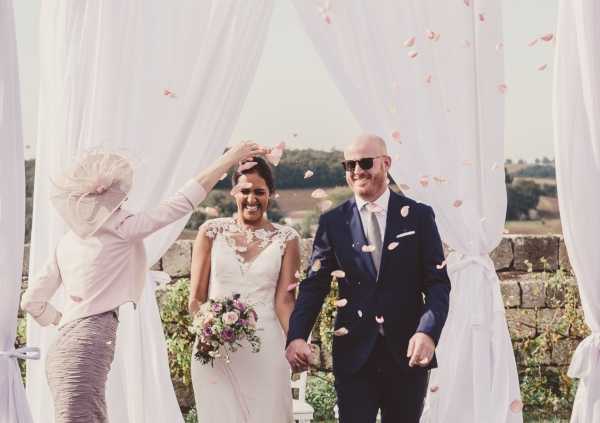 A bride and groom walk back down the aisle as a guest tosses pink rose petals over them during an outdoor ceremony recessional. The ceremony takes place beneath a white draped fabric arch structure, with a countryside or vineyard landscape visible in the background. The bride wears a fitted white gown with lace cap sleeve detailing and carries a loose bouquet of dusty pink and mauve flowers with greenery, while the groom wears a navy suit with a light grey tie and sunglasses. The shot is a medium portrait-style image capturing the couple's joyful expressions mid-confetti toss.
