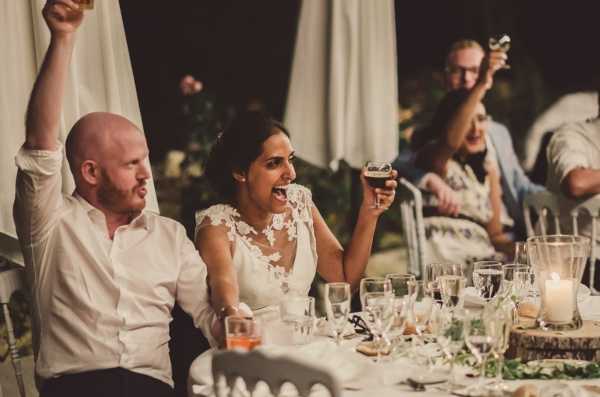 The bride and groom are seated at the head table during an outdoor evening reception, laughing and raising their glasses in a toast. The groom wears a white dress shirt and the bride wears a white lace-appliqué wedding dress with a sheer bodice. The table is set with glassware, candles in glass holders, and low greenery garland centerpieces. Several guests are visible in the background, also raising glasses, in what appears to be a relaxed, informal atmosphere with white draping and warm candlelight. The image is a candid medium shot with a warm, dimly lit nighttime ambiance, consistent with a rustic or boho styling approach.